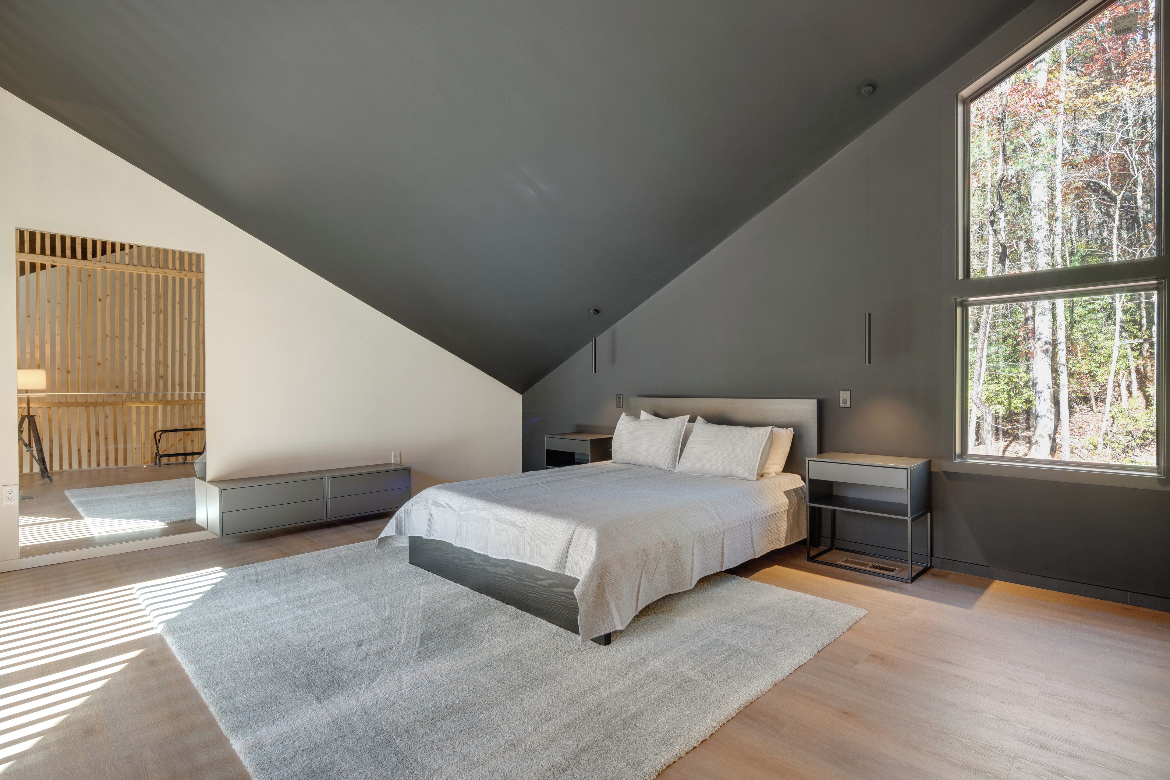 Une chambre moderne avec un plafond en pente, un grand lit, une table de chevet et une fenêtre donnant sur la forêt.