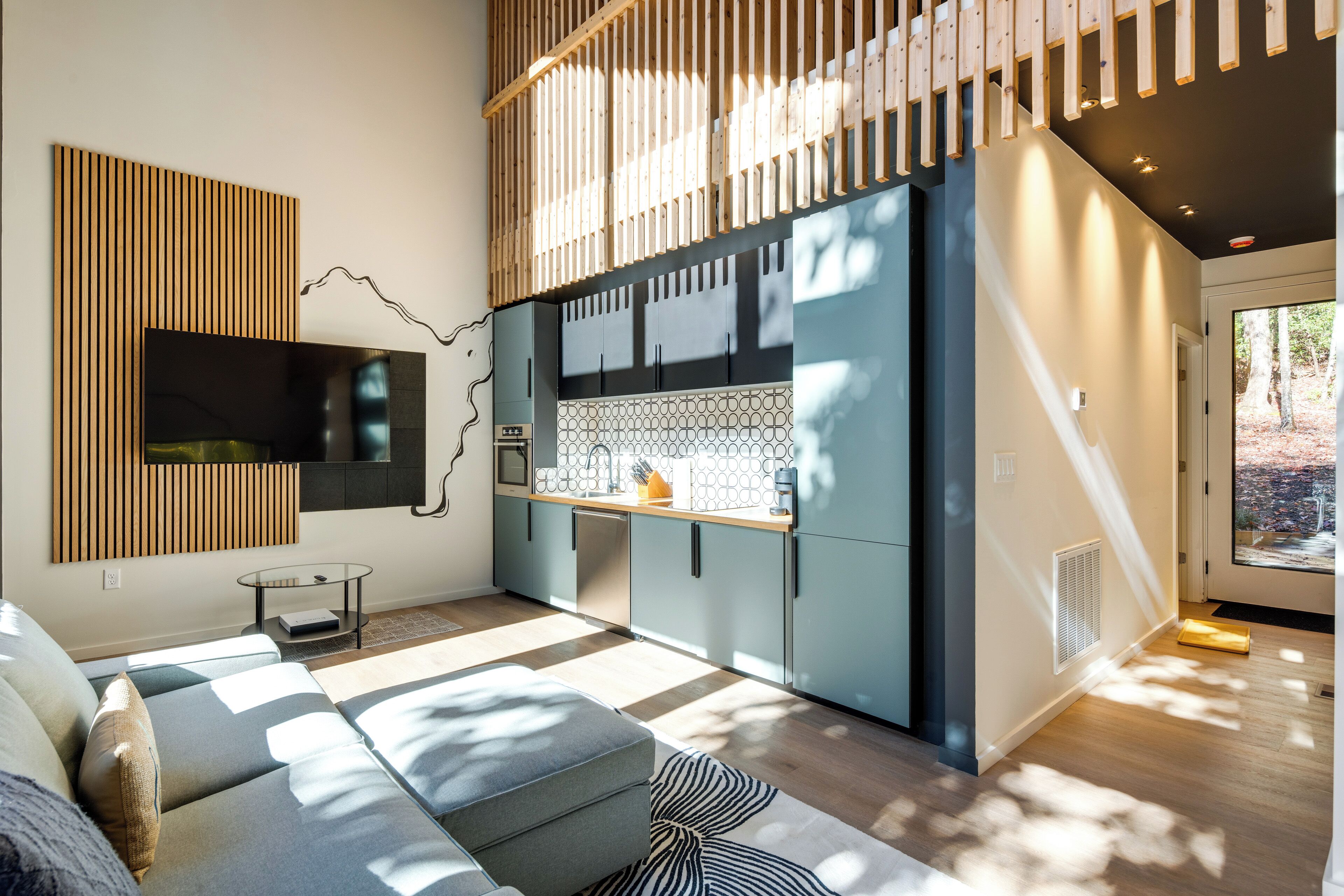 A modern living room with a sofa, a television mounted on a wooden panel, and a kitchen area with cabinets and a countertop.