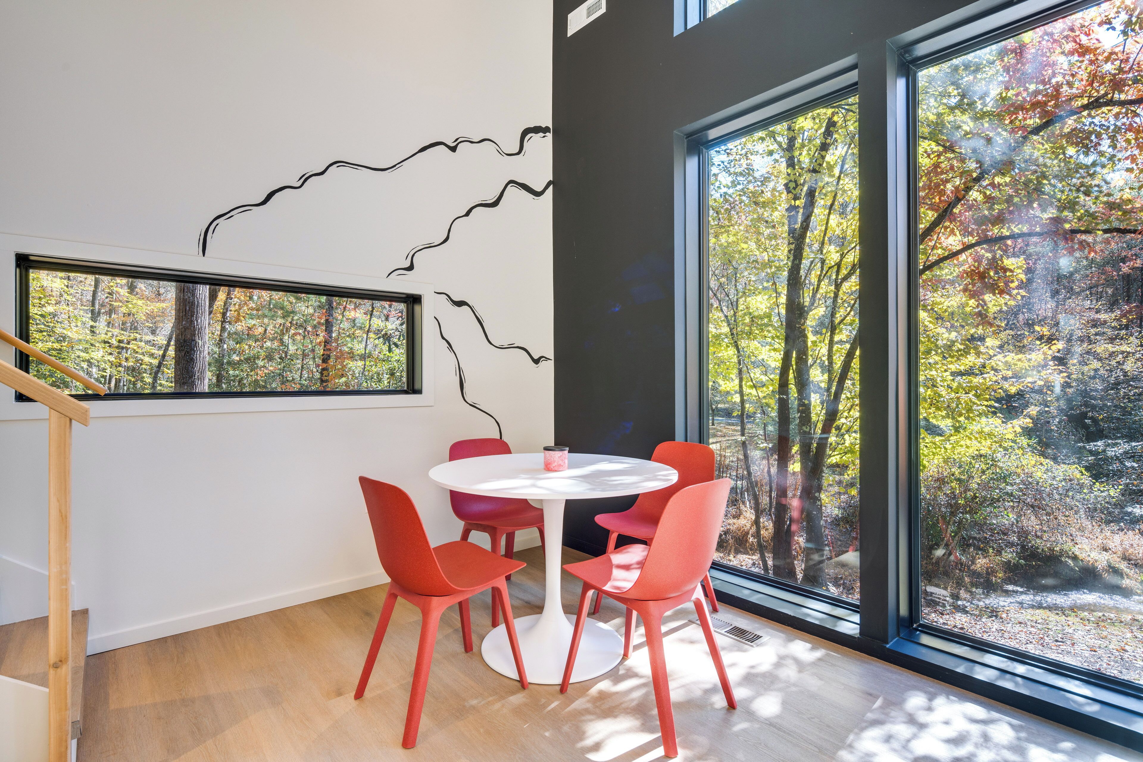 A dining area with a round table and six red chairs, large windows with a view of trees, and a black abstract wall art piece.