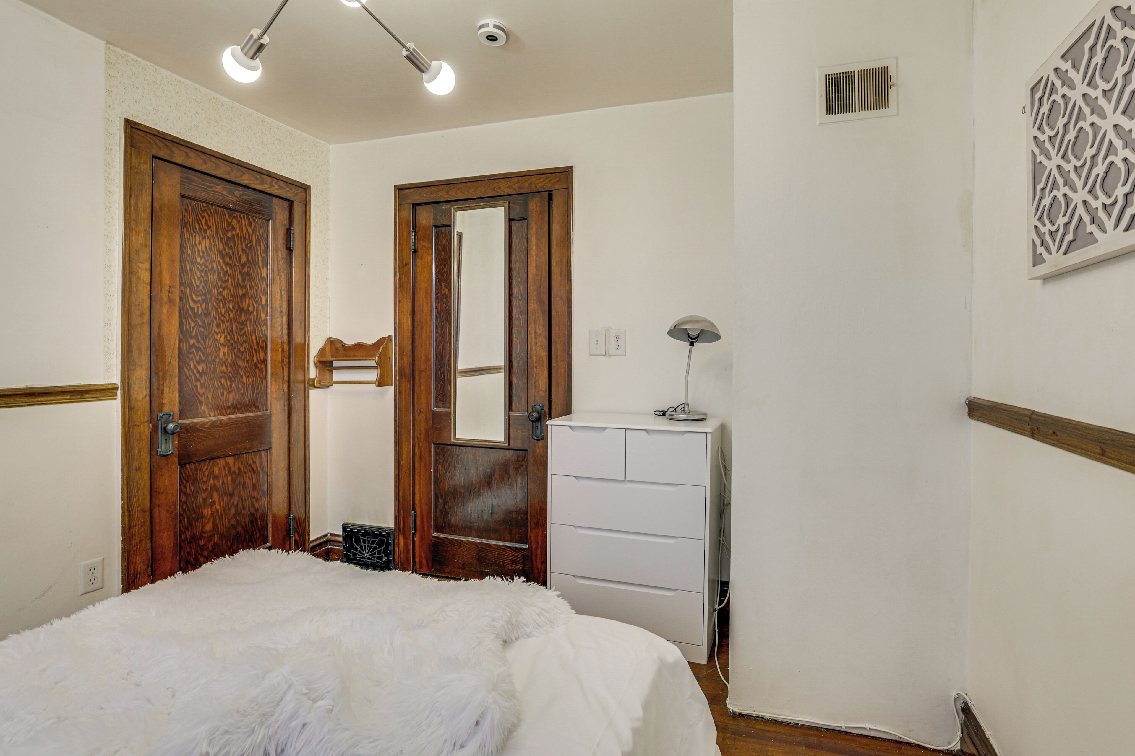 A bedroom with a wooden door, a mirror, a white dresser, and a fluffy white bedspread.