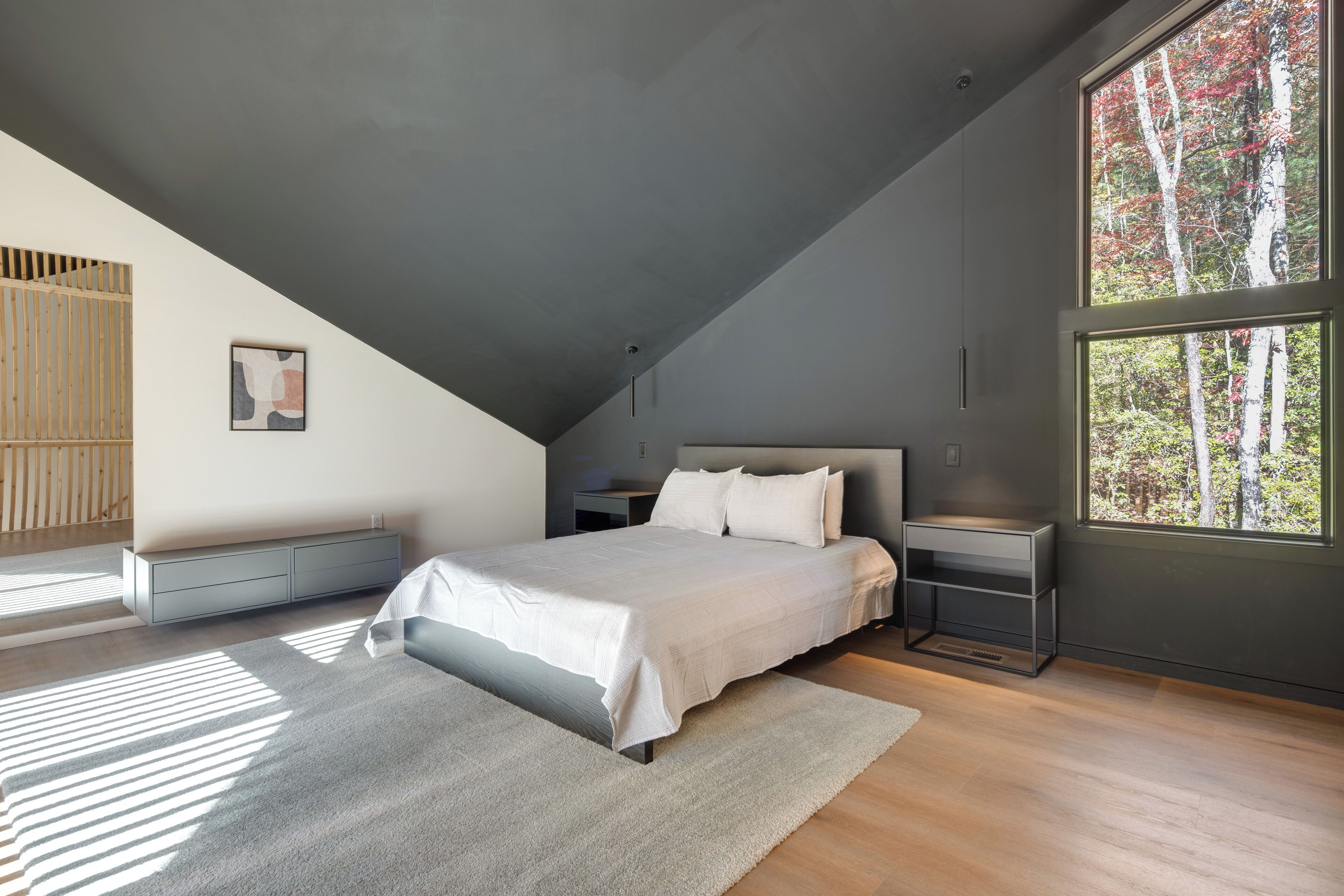 A modern bedroom with a sloped ceiling, a large bed, a bedside table, a nightstand, a window with a view of trees, and a wall-mounted shelf.