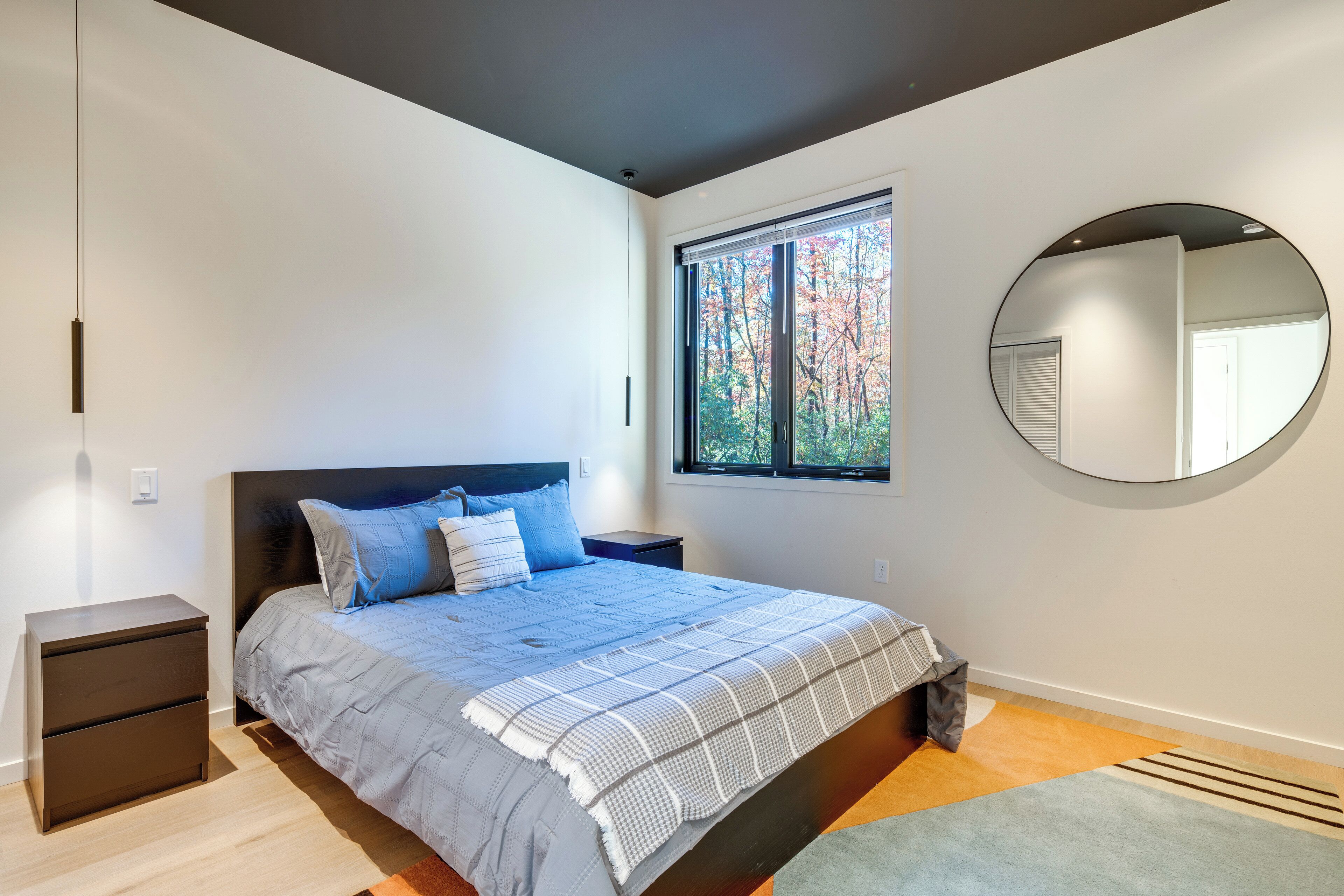 Una camera da letto con un letto, comodini, una finestra vista alberi e uno specchio rotondo appeso al muro.