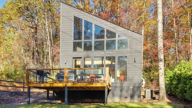Una cabaña moderna de dos pisos con un balcón, ventanas amplias y un paisaje forestal natural.