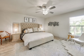 Une chambre à coucher avec un grand lit, un ventilateur de plafond, une table de chevet avec une lampe, une chaise et une fenêtre avec des stores.