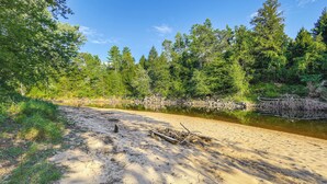 A sandy beach with a calm river, surrounded by dense green forest. - 9 Mi to Hickory Ski Center: Wooded Warrensburg Gem (Warrensburg)
