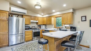 A modern kitchen with wooden cabinets, stainless steel appliances, a central island with bar stools, and a patterned rug. - 9 Mi to Hickory Ski Center: Wooded Warrensburg Gem (Warrensburg)