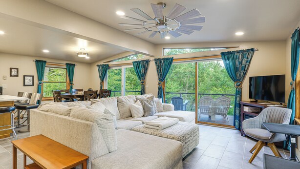 A spacious living room with a sectional sofa, a wooden coffee table, and a TV. There is a dining area with chairs and a table, and large windows with curtains. - 9 Mi to Hickory Ski Center: Wooded Warrensburg Gem (Warrensburg)
