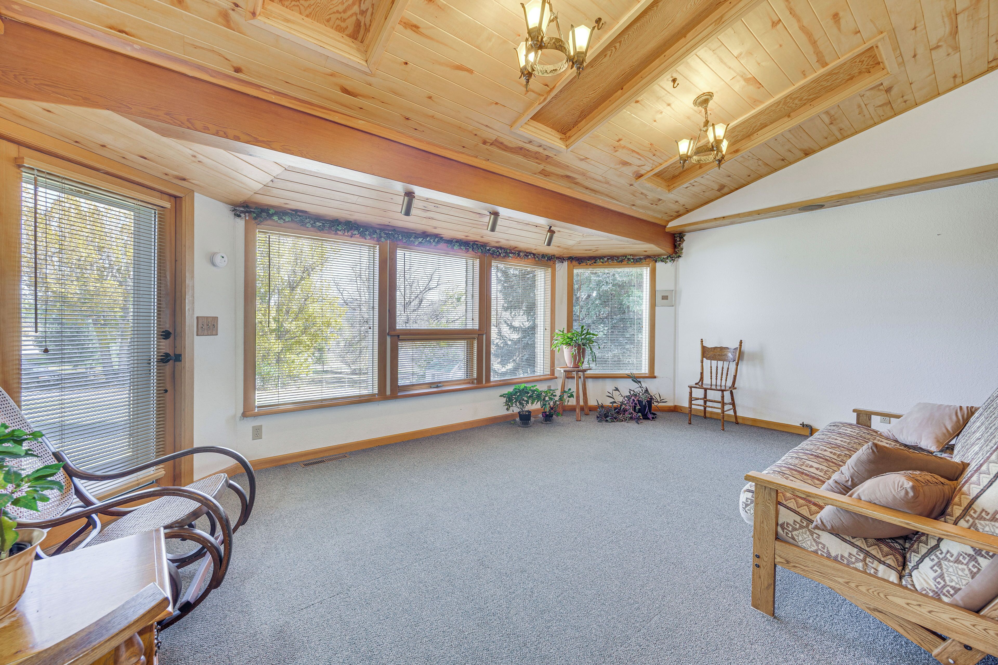 A spacious room with a high ceiling, wooden beams, a large window with blinds, a wooden chair, a sofa, and a small table with a plant.