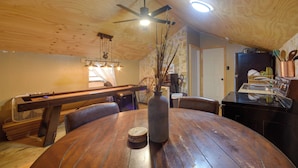 A wooden dining table with a vase and a coaster, a ceiling fan, and a kitchen area with a refrigerator and stove.