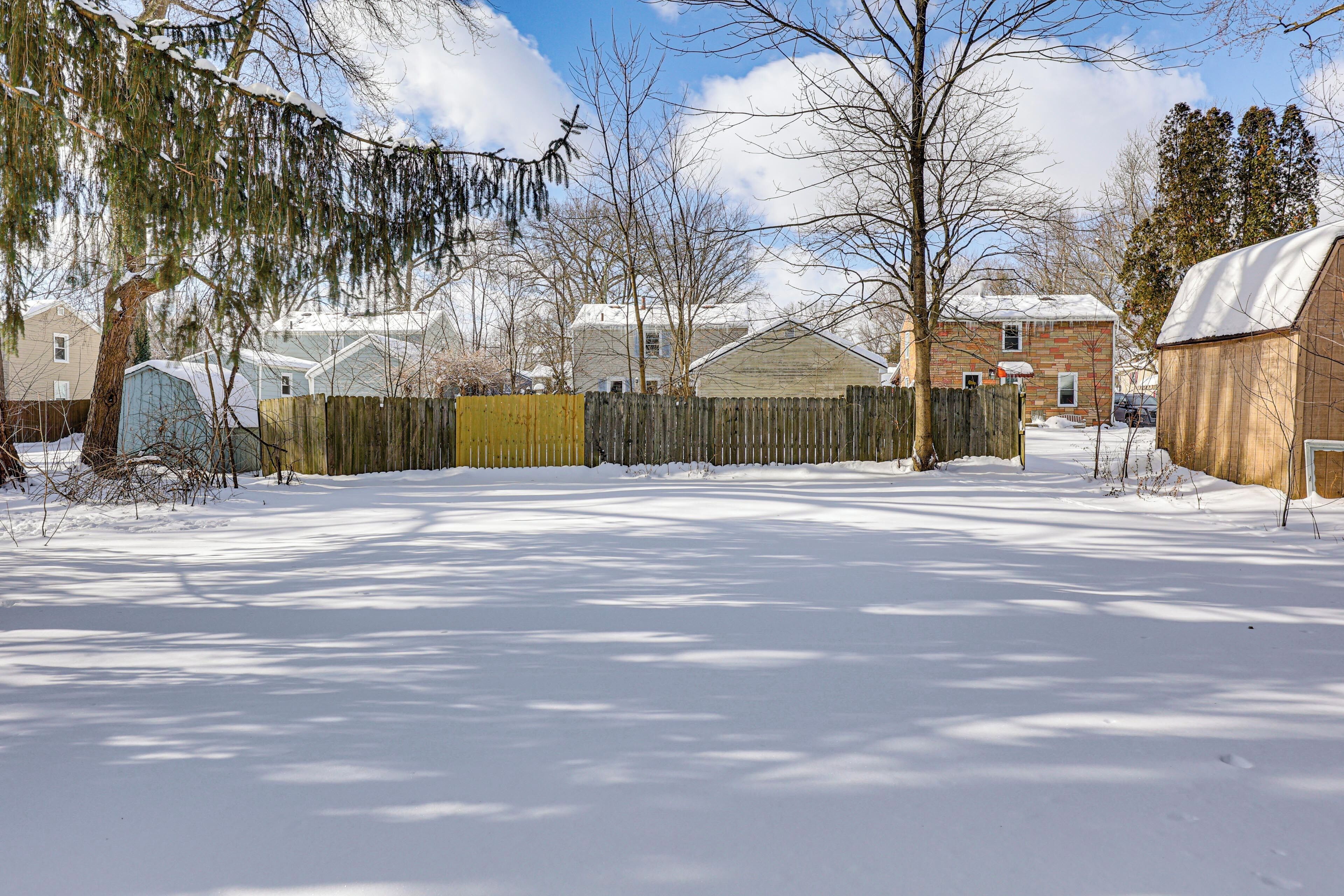 A snow-covered yard with a wooden fence, a shed, and houses in the background.