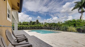 Un área de piscina con solárium empedrado, sillas de descanso y un jardín vallado con árboles.