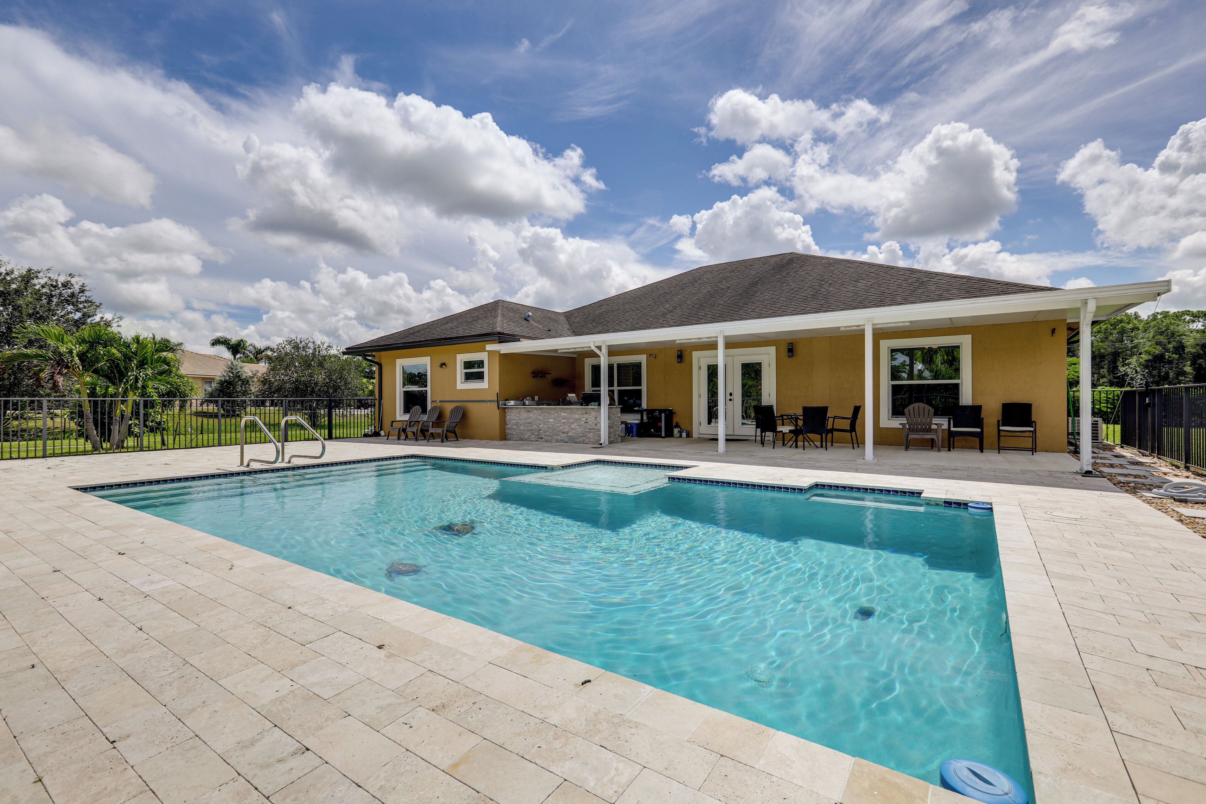 A rectangular swimming pool with a tiled deck, a covered patio area with outdoor furniture, and a house in the background.