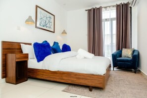 A modern bedroom with a wooden bed, blue and white pillows, a blue chair, and a large window with curtains. - THE LOXWOOD BY EBS LEGEND PROPRETIES (Accra)