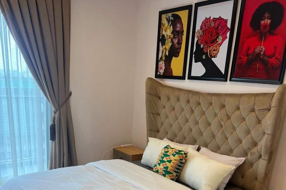 A bedroom with a tufted headboard, a large bed with white bedding, a brown furry rug, and three framed artworks above the bed.