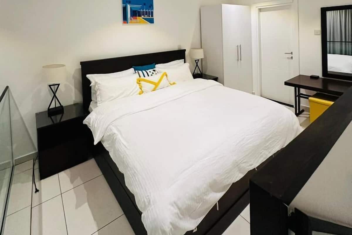 A modern hotel room with a large bed, a desk, a chair, a wardrobe, and a painting on the wall.