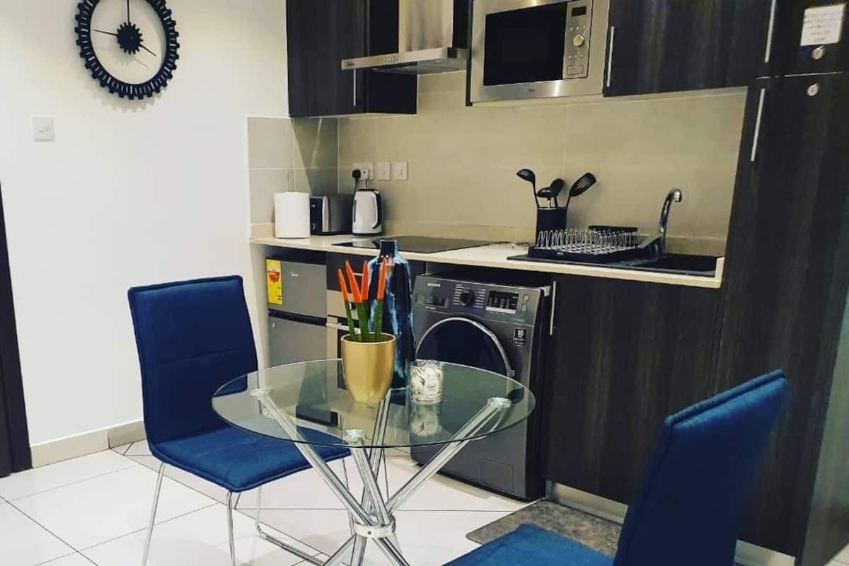 A modern kitchen with a glass dining table, two blue chairs, a microwave, a stove, and a washing machine.