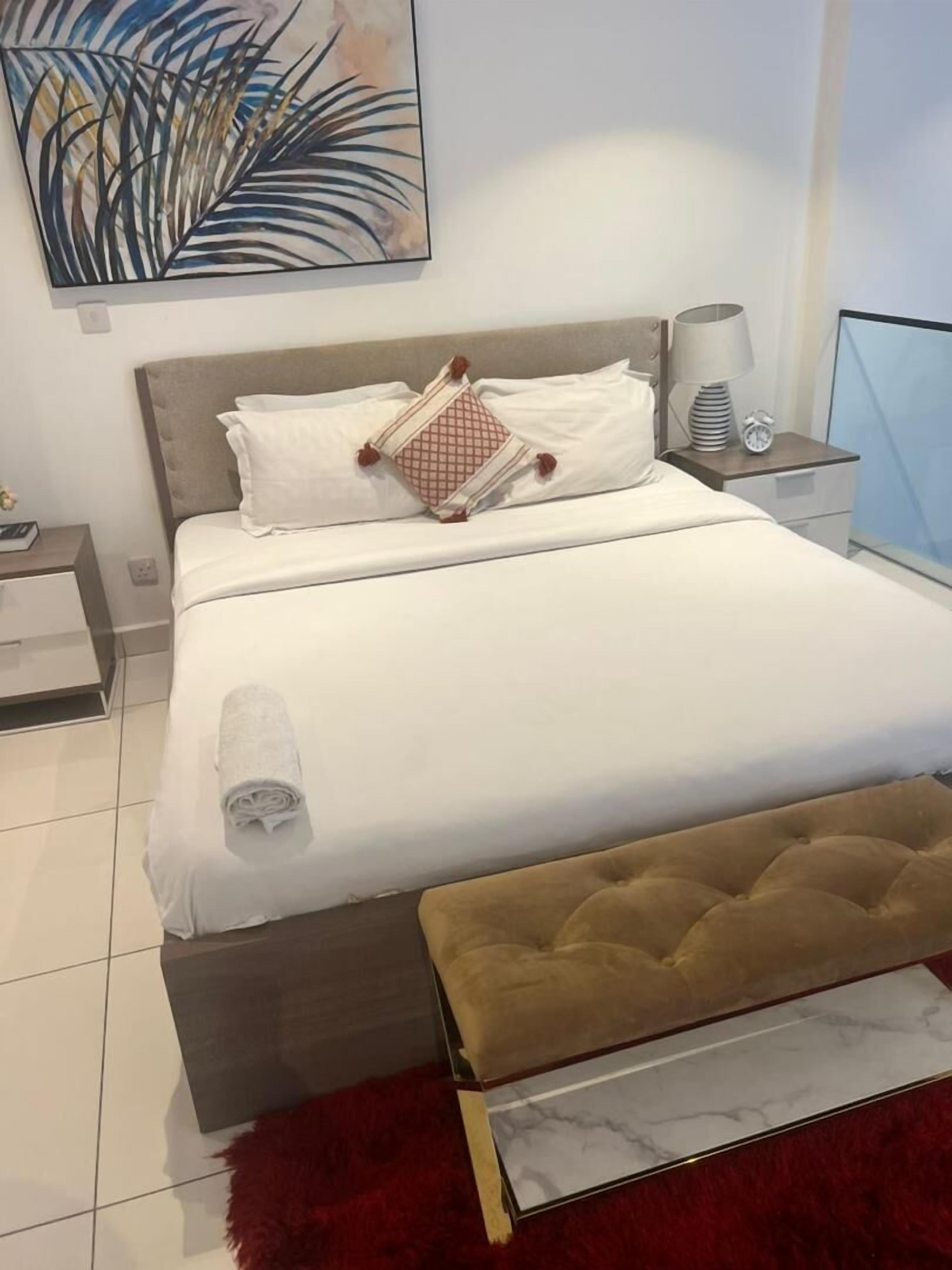 Una cama bien hecha con sábanas blancas y una toalla enrollada, un cabecero capitonado, una mesita de noche con una lámpara, y un cuadro con follaje en la pared.