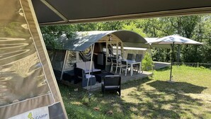 Een kampeerterrein met tenten, een tafel en stoelen onder een overkapping, omringd door bomen.