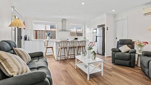 A modern living room with a sofa, armchairs, and a coffee table. A kitchen with a dining area is visible in the background. - Pete's Retreat - Coastal Comfort Just Steps from t (Noraville)