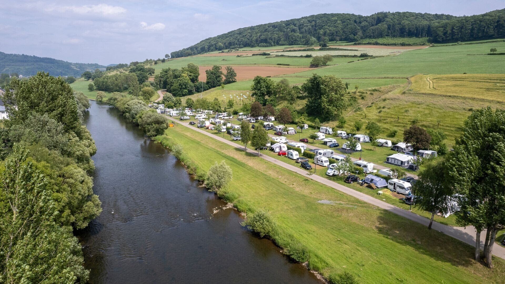 A caravan park with rows of caravans along a river, surrounded by green fields and trees.