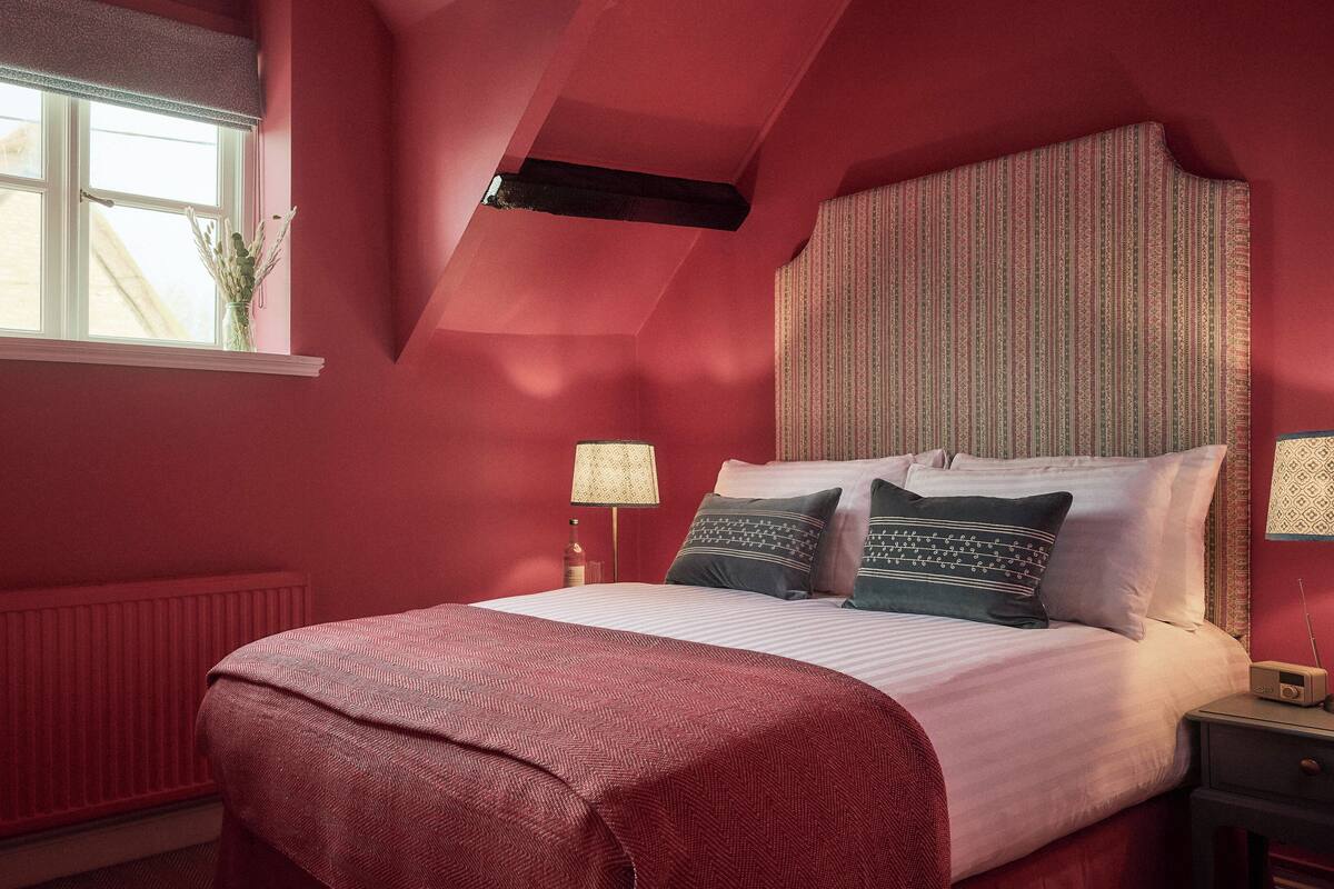 A bedroom with a sloped ceiling, a bed with patterned bedding, two bedside lamps, and a window with a view of a building outside.