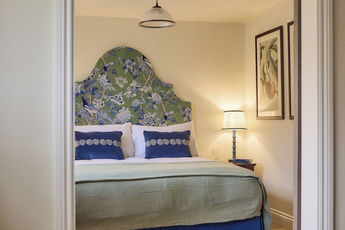 A bedroom with a floral headboard, a bed with blue bedding, a nightstand with a lamp, and two framed pictures on the wall.