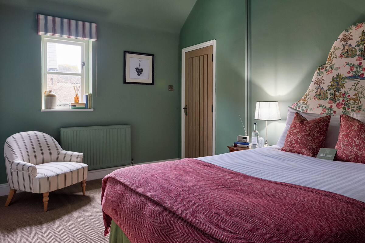 A bedroom with a bed, a chair, a radiator, a door, a window, and a wall with a framed picture.