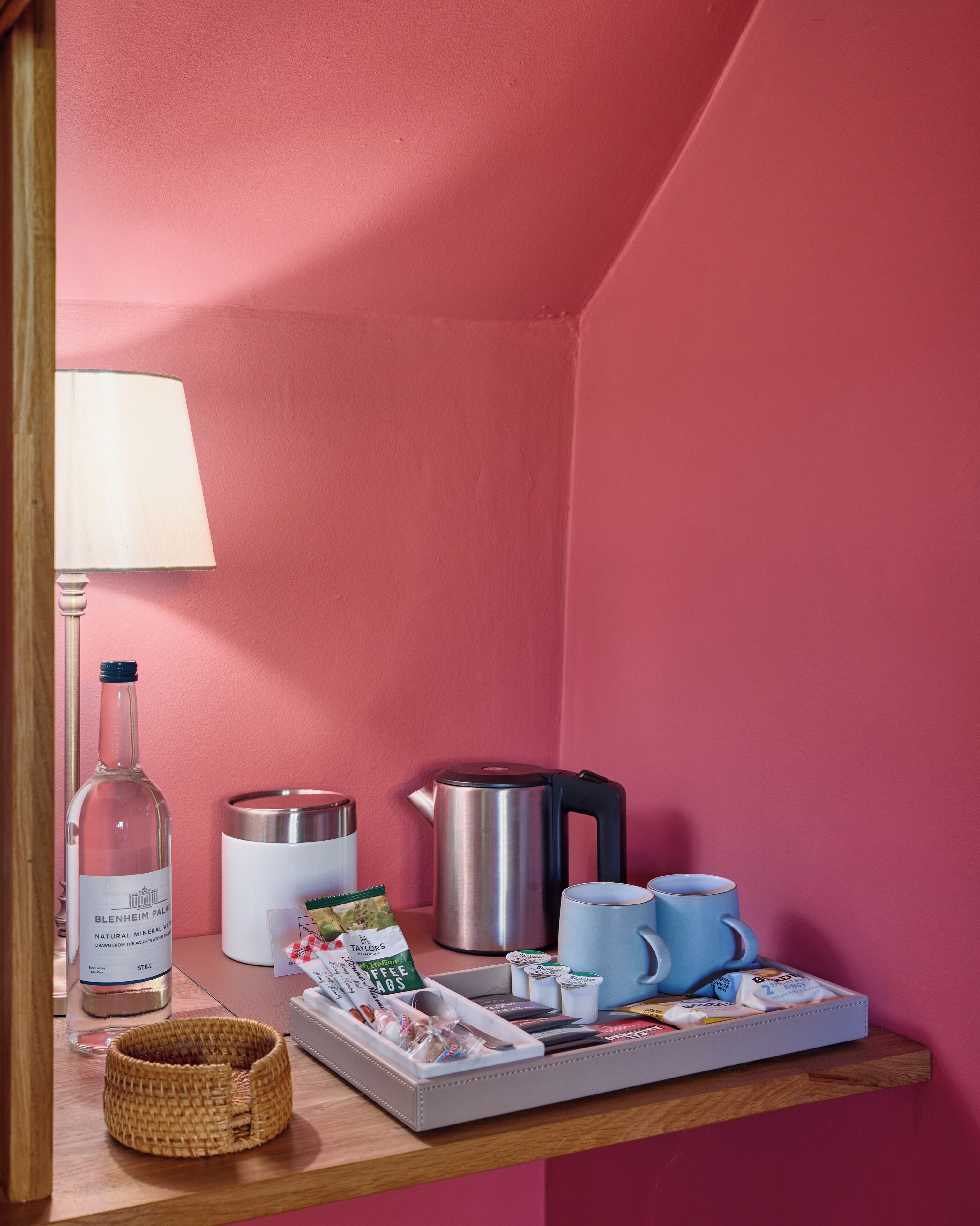 A pink-walled room with a wooden shelf holding a bottle, a kettle, a coffee maker, a mug, and a tray with coffee pods and a spoon.