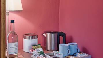 A pink-walled room with a wooden shelf holding a bottle, a kettle, a coffee maker, a mug, and a tray with coffee pods and a spoon.