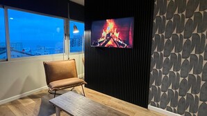 A modern living room with a large TV displaying a fireplace, a leather chair, a wooden table, and a view of the city through large windows. - Design apartment in historic district with river view (Montevideo)