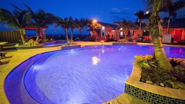A swimming pool with a curved edge, surrounded by palm trees and a building with red lighting. - The Rod and Reel Resort-Suite 5 (Anna Maria)