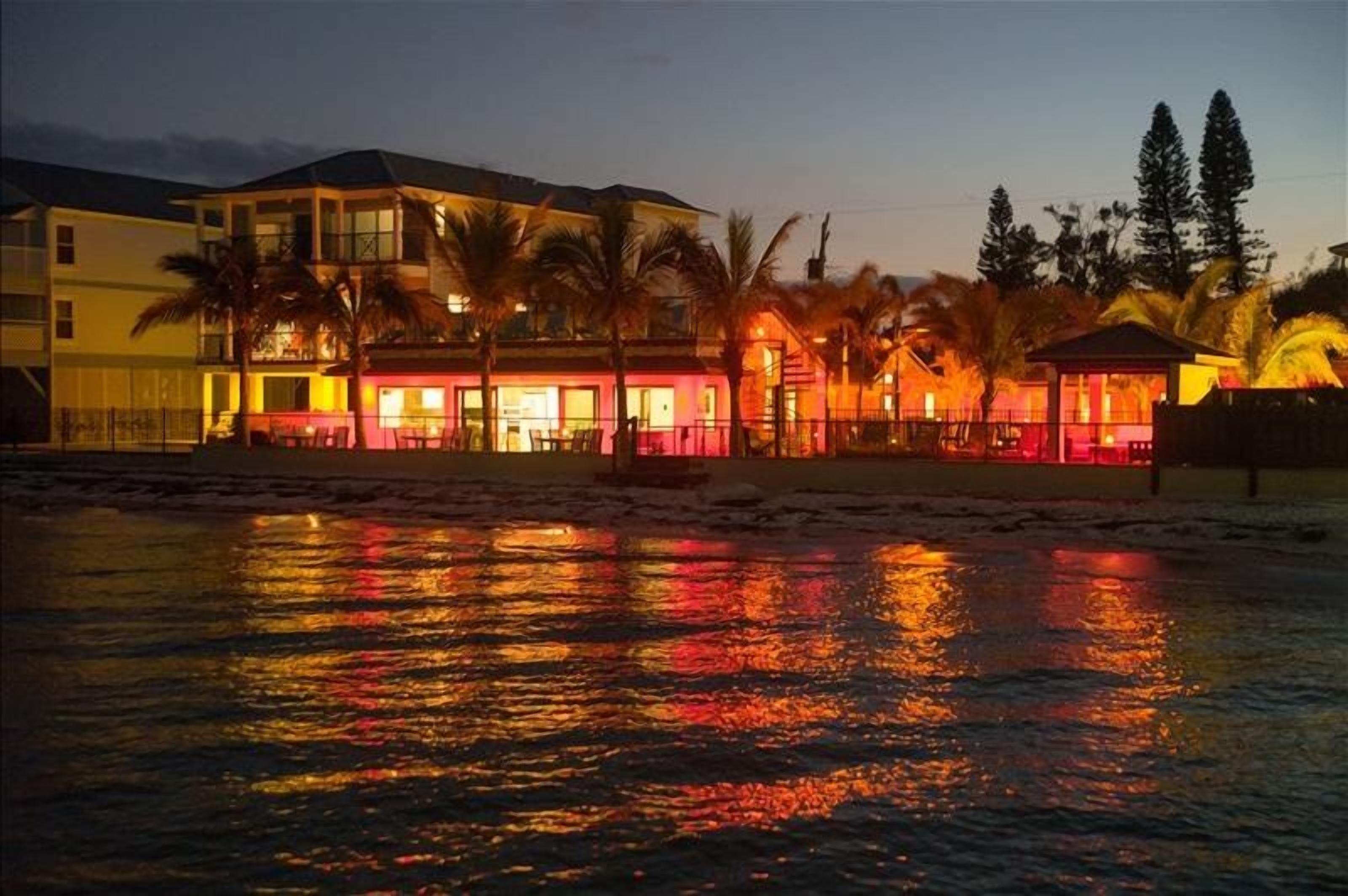 Ein Strandhotel mit mehreren Gebäuden, Palmen und einer spiegelnden Wasseroberfläche.
