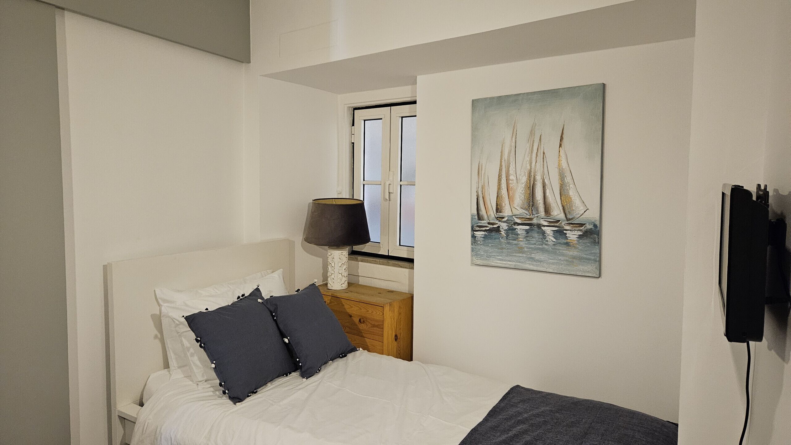 A bedroom with a bed, a nightstand with a lamp, a window, and a painting of sailboats.