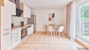 A modern kitchen with white cabinets, a stainless steel refrigerator, and a dining area with a round table and white chairs. - Lena Your break (Buchen)