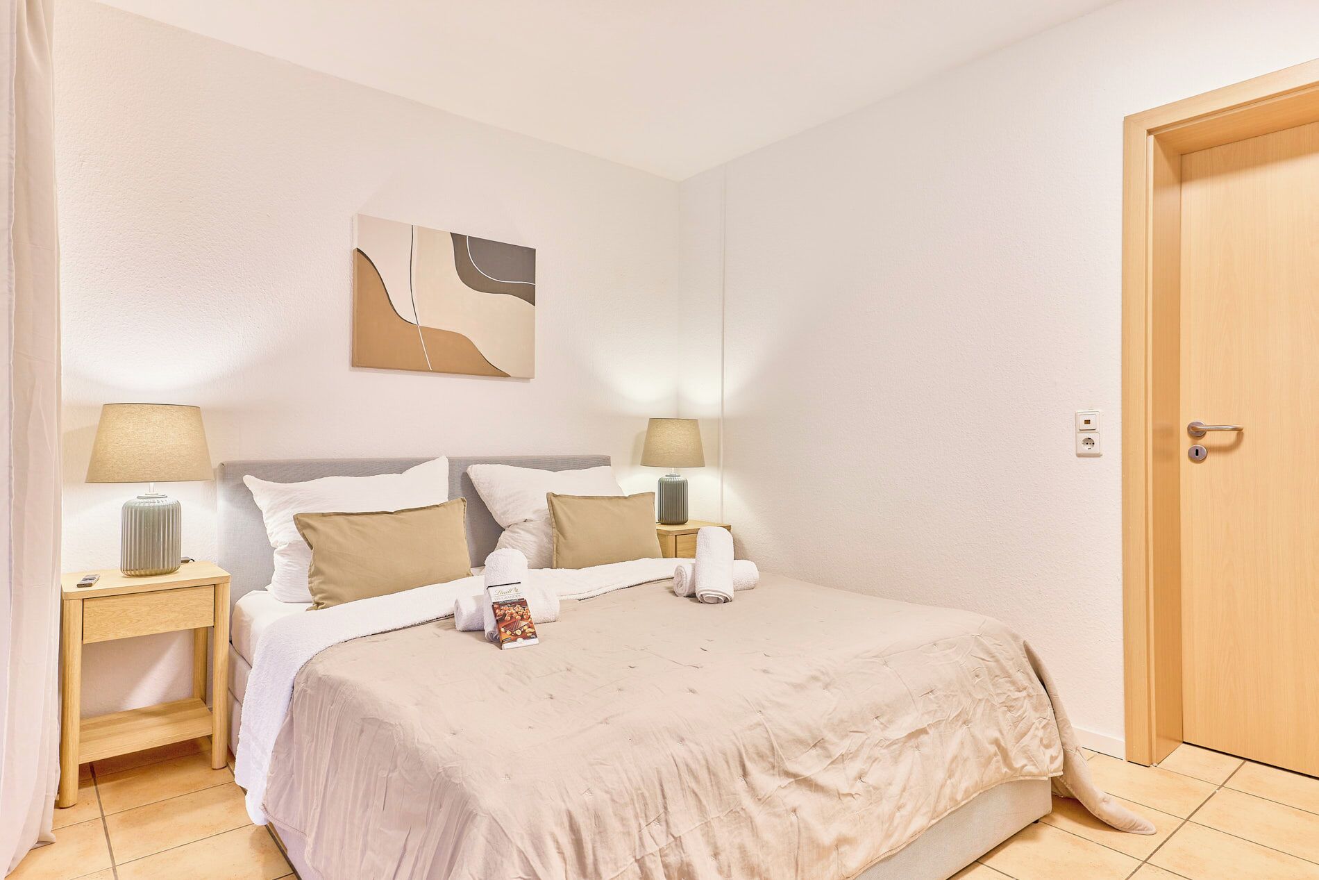 A neatly made bed with a beige bedspread and pillows, two bedside lamps, a wall art piece, and a wooden door.
