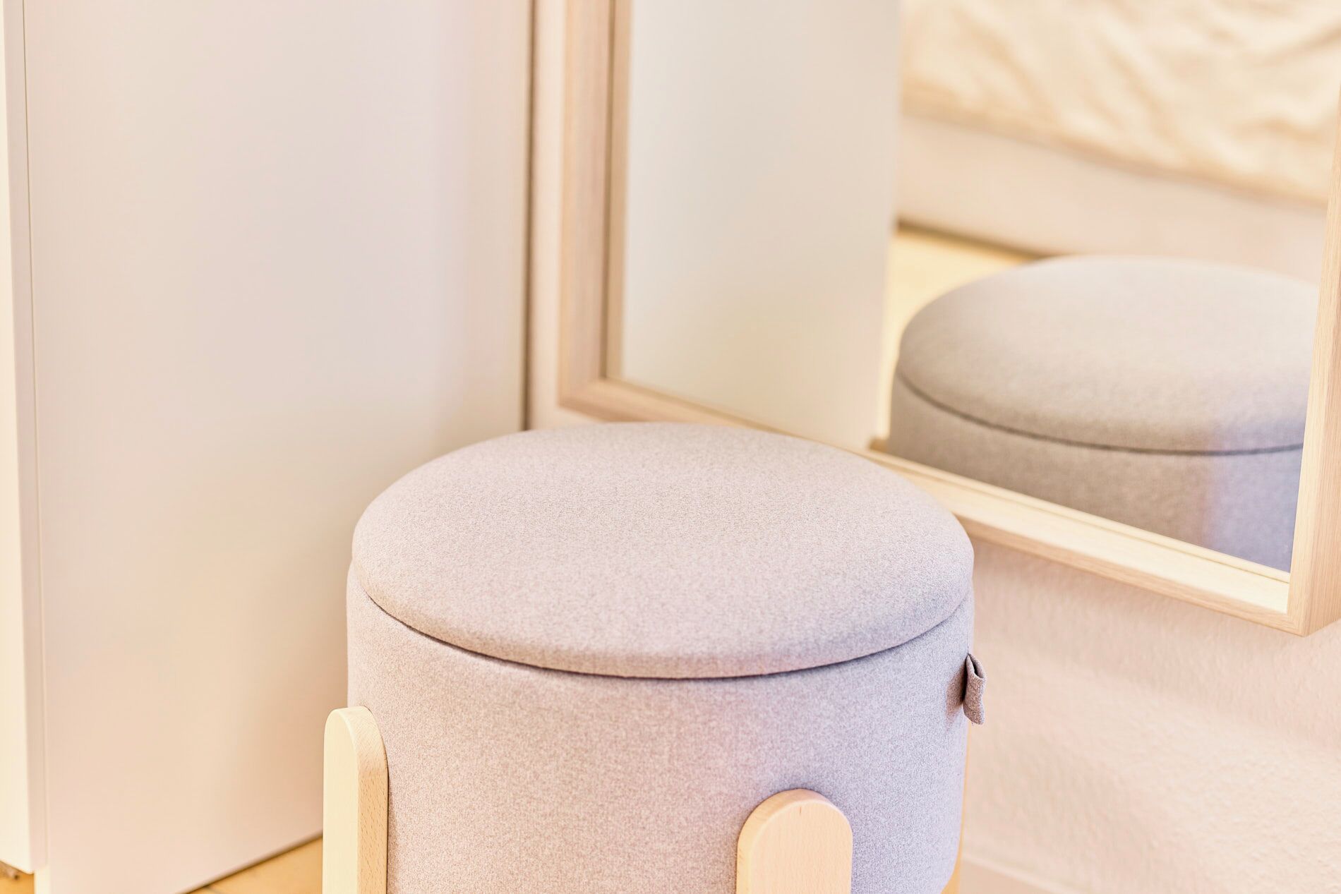 Un pouf rotondo, di colore chiaro, con base in legno, posizionato di fronte a uno specchio che riflette un letto.