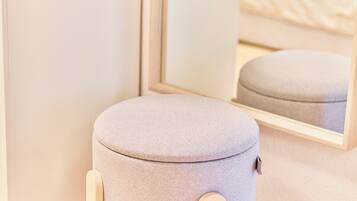Un pouf rotondo, di colore chiaro, con base in legno, posizionato di fronte a uno specchio che riflette un letto.