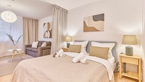 A modern hotel room with a large bed, two bedside tables with lamps, a sofa, and a small side table with a vase. - Lena Your break (Buchen)