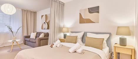 Quarto de hotel moderno com uma cama grande, duas mesas de cabeceira com luminárias, um sofá e uma mesinha lateral com um vaso.