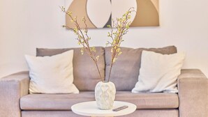 A living room with a beige sofa, two white cushions, a white vase with branches, and a remote control on a round table. - Lena Your break (Buchen)
