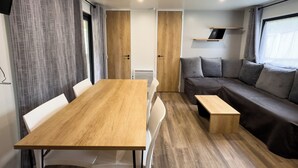 A compact living space with a wooden table, a sofa, and a bench. There are curtains, a door, and a shelf with a book. - Domaine Des Acacias Tours (La Ville-aux-Dames)