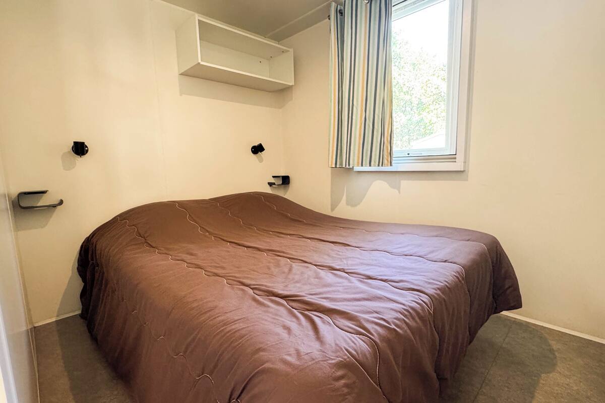 A bedroom with a single bed, a window with striped curtains, a wall-mounted shelf, and a door with a handle.