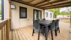 A covered outdoor deck with a wooden table and chairs, a sliding glass door, and a view of a mobile home in the background. - Domaine Des Acacias Tours (La Ville-aux-Dames)