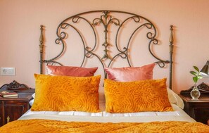 Un lit avec une tête de lit en métal, deux oreillers, une table de chevet avec une lampe et une petite plante.