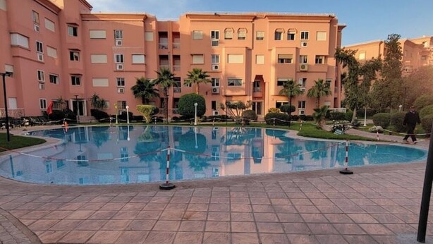 Ein Schwimmbad, umgeben von einer gefliesten Terrasse, mit einem mehrstöckigen Gebäude im Hintergrund.