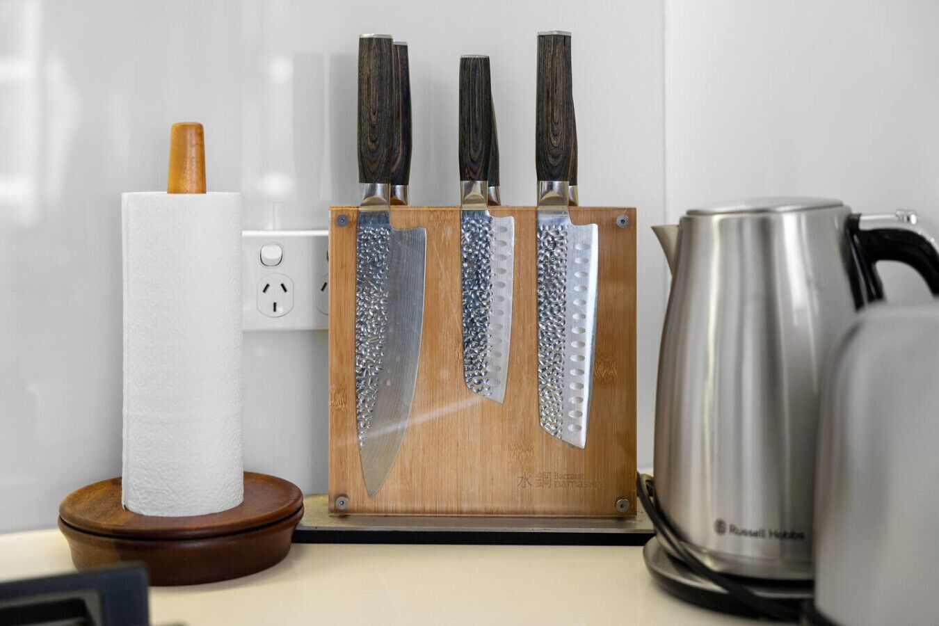 A kitchen counter with a wooden knife block holding three knives, a paper towel holder, and a kettle.