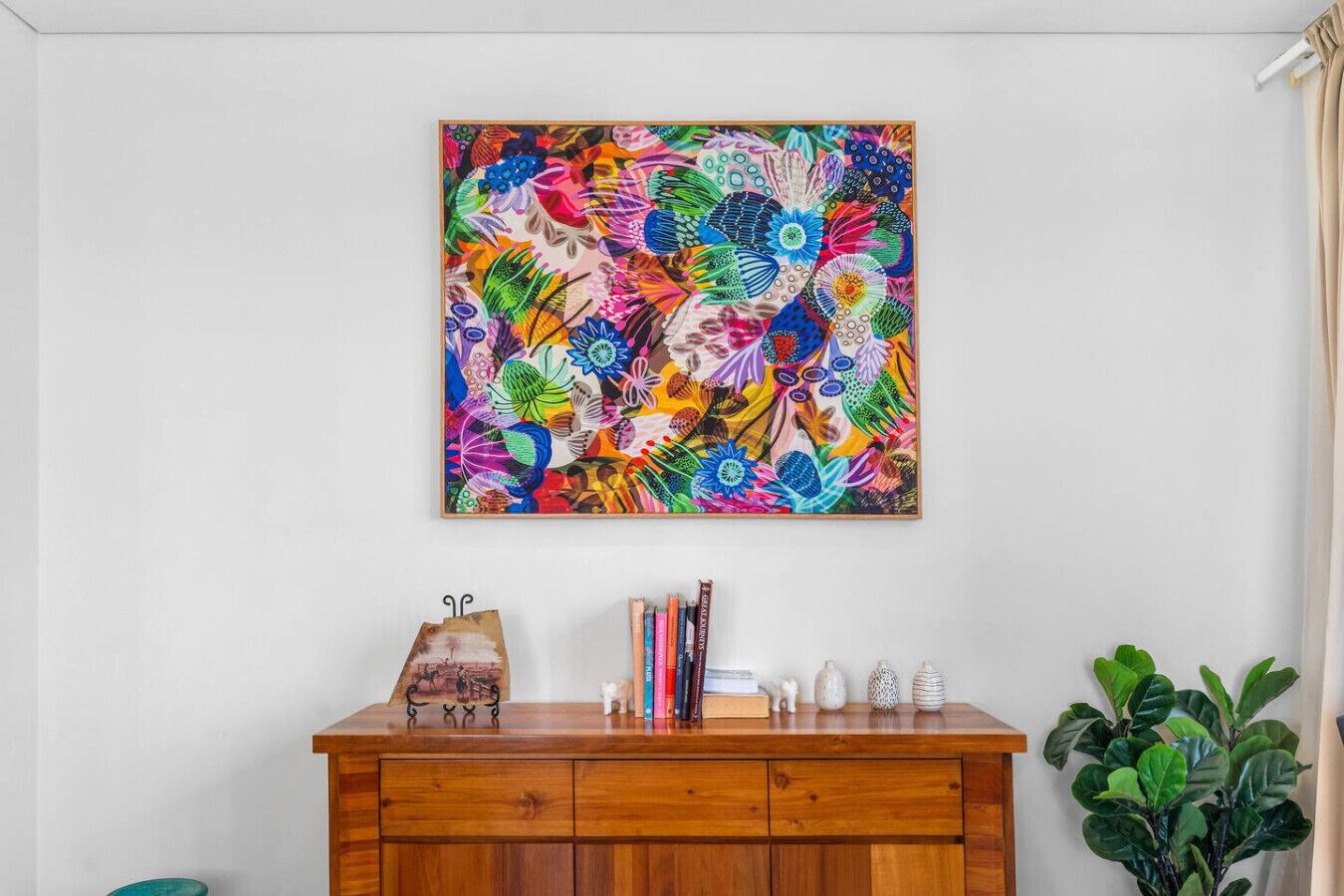 A colorful abstract painting on a wall, a wooden cabinet with various items, and a green plant.