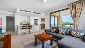 A modern living room with a grey sofa, wooden coffee table, and a colorful rug. A kitchen with white cabinets and a dining area with two bar stools is visible in the background. - Zen Paradiso Harbour View Pool Balcony (Darwin)