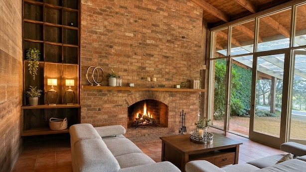 A cozy living room with a brick fireplace, a sofa set, a wooden shelf with decorative items, and a large window offering a view of greenery. - Country Homestead in Avenel (Avenel)