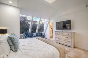 Ein Schlafzimmer mit einem großen Bett, einem Fernseher, einer Kommode und Blick ins Freie durch große Fenster.